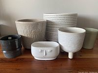 Lot of seven variously sized and styled flower and plant pots arranged in one row on a wooden surface against a light-colored wall