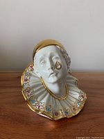 Front view of white porcelain Pierrot clown bust figurine with 24K gold cap and collar edges, Swarovski teardrop crystal below eye, and pink and blue floral details on ruffled collar.