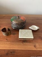 Four vintage jewelry-related items on a wooden surface: painted metal jewelry dish with lid, white mini dish, fabric-covered jewelry travel case closed, brass bangle.