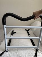 Front view of safety bed rail with black padded handle, three horizontal white bars, and base straps extending under mattress.