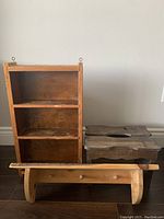Wooden shelf with three compartments in medium brown finish beside a small carved wooden stool and a lighter wooden shelf with hanging pegs.