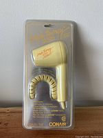 Full front view of vintage baby yellow Conair hair dryer in clear plastic blister packaging, showing branding and coiled cord.