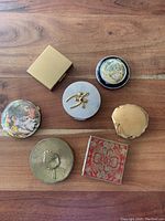 Top view of seven vintage compacts showing a variety of shapes, patterns, and finishes including artwork and metallic designs.