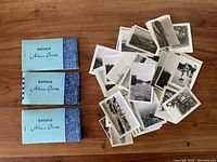 Three Eaton's photo albums with blue covers and decorative pattern stacked on wooden surface, along with loose black and white photos scattered next to them.