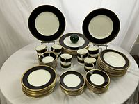 Overview of stacks of matching black-and-gold rimmed plates, bowls and cups on white tablecloth