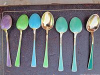 Seven teaspoons laid out on dark surface showing enamel colors and gilt interiors