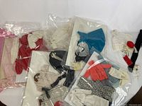 Multiple sealed plastic sleeve packs containing various doll clothing items: capes, coats, skirts, sweaters, and accessories