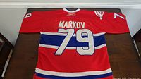 Full back view of the hockey jersey laid flat showing red color with white and blue stripes, Markov name and number 79.