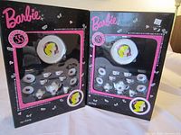Photo of two Barbie 35th Anniversary tea sets in original black packaging with pink and white designs, showing tea utensils through clear window.