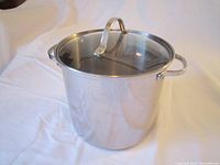 Front and side view of a polished stainless steel pot with transparent lid placed on a white surface.