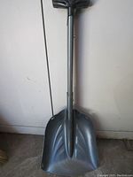 Full view of the Black Diamond emergency shovel showing the broad, metallic blade, shaft, and ergonomic T-grip handle, positioned upright against a wall.