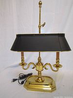 Front view showing the brass base with two curved arms, candle-like light fixtures, black power cord, and black rectangular metal shade with vertical adjustment rod and finial.