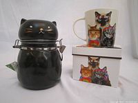 Photo showing both items together: a black ceramic cat storage jar and a colorful cat-themed ceramic mug with its box.
