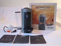 Photo showing DeLonghi Nespresso Vertuo Plus espresso machine, power cord, clear water tank, original box, and instruction manuals on flat surface against white background.