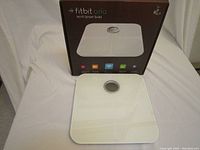 White Fitbit Aria Wi-Fi Smart Scale with circular digital display, placed on a white cloth background, with original box standing in the background