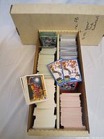 Top view of a large cardboard box divided into four compartments full of coins sorted by series and sport, showing cards from 1989 Score Football and 1991-1992 Upper Deck Basketball with example card front images visible.