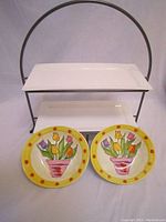 Black metal stand holding two white rectangular ceramic trays with two colorful plates in front showing potted tulip design.