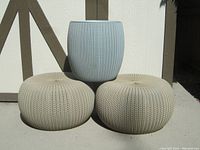 Three pieces displayed outdoors, two beige hassocks and one blue planter stand