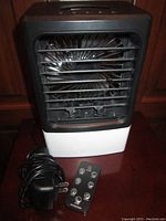 Front view of the portable cooling fan showing black front grill, white base, and fan blades. AC adapter plug and black remote control placed beside unit.