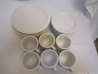 Top view showing stacked white dinner plates, salad plates, and arranged mugs on white backdrop.