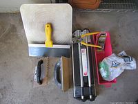 Overview of tile tools including a manual tile cutter, notched and flat trowels, grout spreader, tile nippers, and a pink container with accessories.