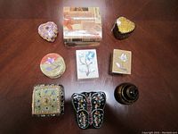 Top view of 9 assorted trinket boxes arranged on table, showing various shapes and designs including stone floral inlays and lacquered wood with metallic details.