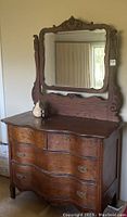 Full view of antique wooden dresser with attached beveled mirror showing curved drawer fronts, dark wood, and decorative carving on mirror and legs.