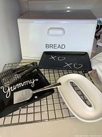 Photo of decorative black serving plates, cooling rack, white ceramic dish shaped like an oval handle, and spatula with background white metal bread box.