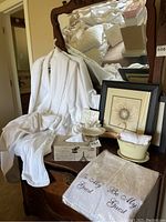 Wide view of the lot showing white robes on wooden chair, framed pictures behind, tissue box, ceramic accents, and towel package.