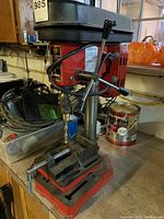 Front view of the Brico drill press showing the entire tool with clamping vice, power cord, and workspace.