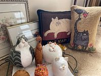 Display of two hand-hooked wool pillows with cat designs, several cat figurines, and two framed pictures in the background.