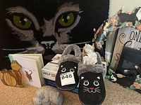 Photo showing black door mat with cat eyes design, black slippers with cat faces, wooden cat decor, and a metal wreath with painted cats.