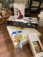 Multiple boxed kitchen tools including KitchenAid rotary grater, OXO mandoline slicer, marble cheese plate, Trudeau cheese shaver, and cheese markers in packaging