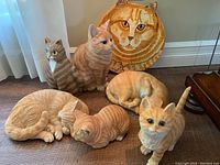 Group of seven cat figurines in orange, brown, and tan hues, empty area behind includes white curtains and beige wall