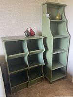 Two green wooden shelving units side by side. Taller unit with four shelves on right, shorter unit with six compartments on left. Curved side design visible, pears on top as props.