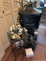View of the black metal mailbox on tall stand with plant stand holding artificial white flowers next to it, artificial greenery draped.