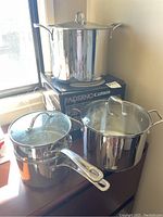 Set of three stainless steel pots on a windowsill including a large 12L pot on its box and two smaller pots with lids