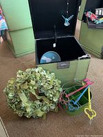 Open green wooden storage box with black interior, a ceramic plate inside, colorful metal key-shaped items organized beside it, and a floral wreath with green leaves and ribbons.