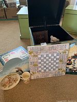 Green Pier 1 storage box with lid open and assorted items displayed in front including a small ceramic plate, a ceramic cup, decorative chess/checkerboard, teddy bear figurine, and a wooden country clatch figurine.