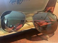 Close-up of heavier metal frame Maui Jim sunglasses with dark lenses showing dust and reflections. Case lid with Maui Jim logo in background.
