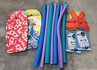 Four boogie boards, seven pool noodles, and an orange life jacket are laid out flat on a concrete floor. The boogie boards have different covers and designs, pool noodles are foam and colorful, and life jacket is secured with straps.
