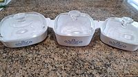 Front view of three white Corningware Cornflower casserole dishes with lids arranged side by side on countertop showing the blue cornflower pattern.