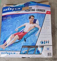 Front of the Kelsyus Deluxe Floating Lounger box showing man reclining on the blue inflatable pool lounger with branding and product details.