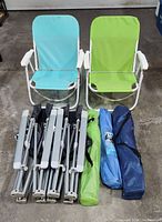 Two beach chairs, one blue and one green, with white frames and armrests, alongside five folded lawn chairs packed in carrying bags.
