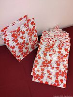 Stack and single view of large gift bags with overlapping red and orange hearts pattern, white rope handles, and matching tags, shown against red surface and white wall.