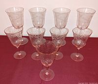 Nine pink Depression glass stemware pieces arranged on a maroon background, showing different shapes and etched floral designs.