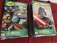 Multiple sealed Crayola giant coloring books with Teenage Mutant Ninja Turtles and Star Wars (Darth Vader) covers arranged on red fabric surface