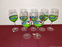 Seven hand-painted stemware glasses with Cape Cod Nauset scenes showing lighthouses, houses, and greenery from front angle.
