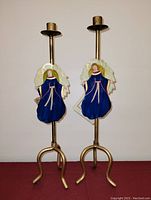 Pair of vintage gold-tone metal candlesticks each with an angel decoration in blue dress and yellow wings, standing on tripod bases.