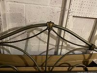 Close-up image of metal headboard featuring crown detail and twisted metal design, in light antique brass/bronze finish.